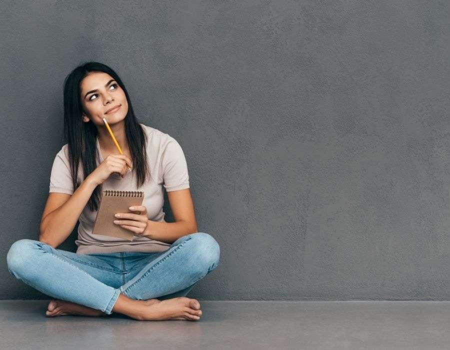 woman-sitting-crosslegged-against-grey-wall-tshirt-blue-jeans-long-dark-hair-holding-notepad-pencil-to-side-of-face-thoughtful-expression