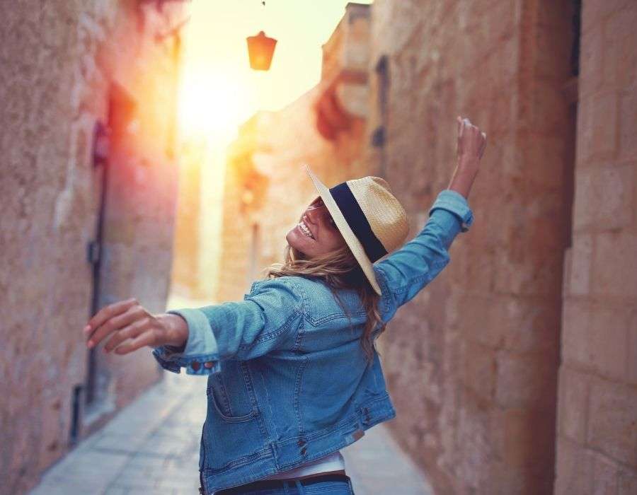 woman-blonde-hair-denim-jacket-straw-hat-walking-down-old-stone-street-arms-outstretched-smiling-chasing-your-dreams-blog-post