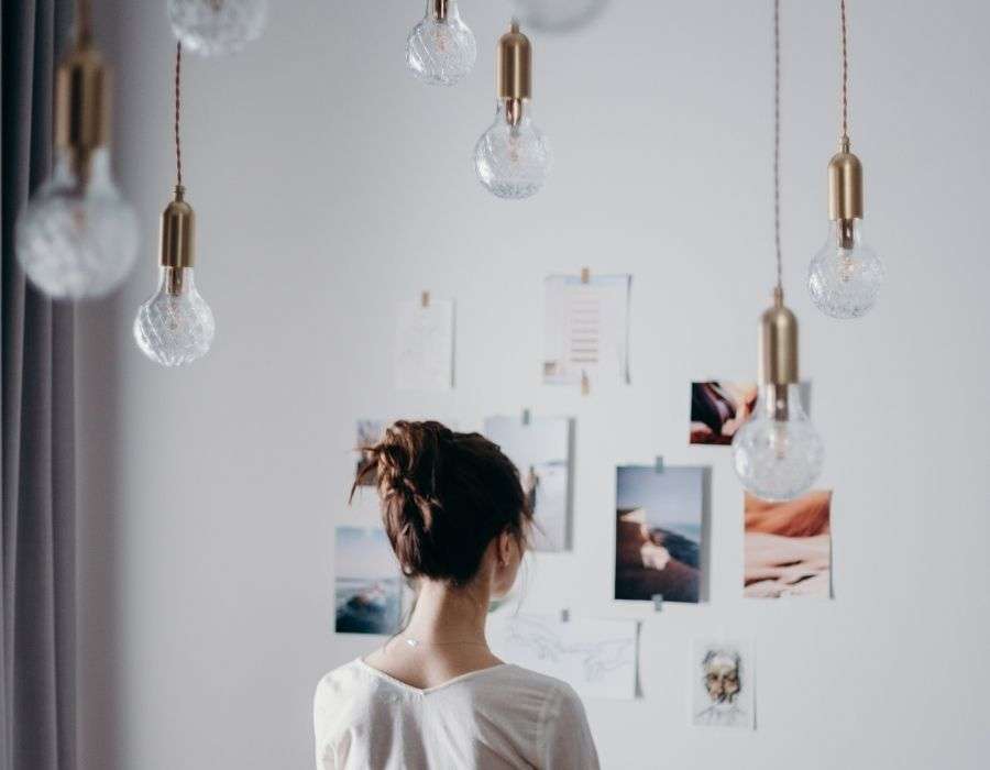 woman-from-behind-brown-hair-in-bun-staring-at-collage-of-pictures-on-wall-for-inspiration-multiple-dangling-lights-hanging-from-ceiling