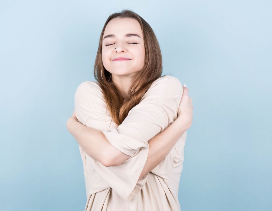 self-love-blog-woman-smiling-arms-wrapped-around-herself-blue-background